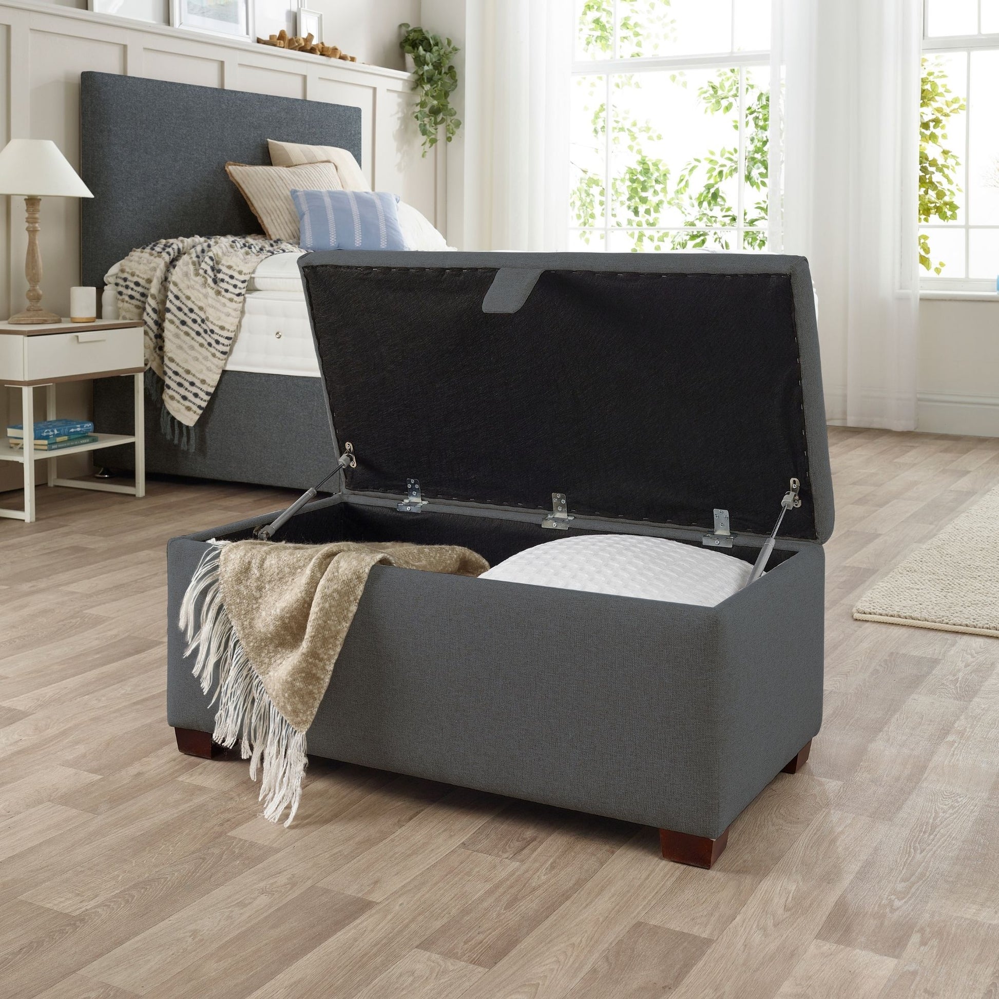 The Woolroom Ottoman Storage Box in dark grey upholstery stores pillows and throws and other items.