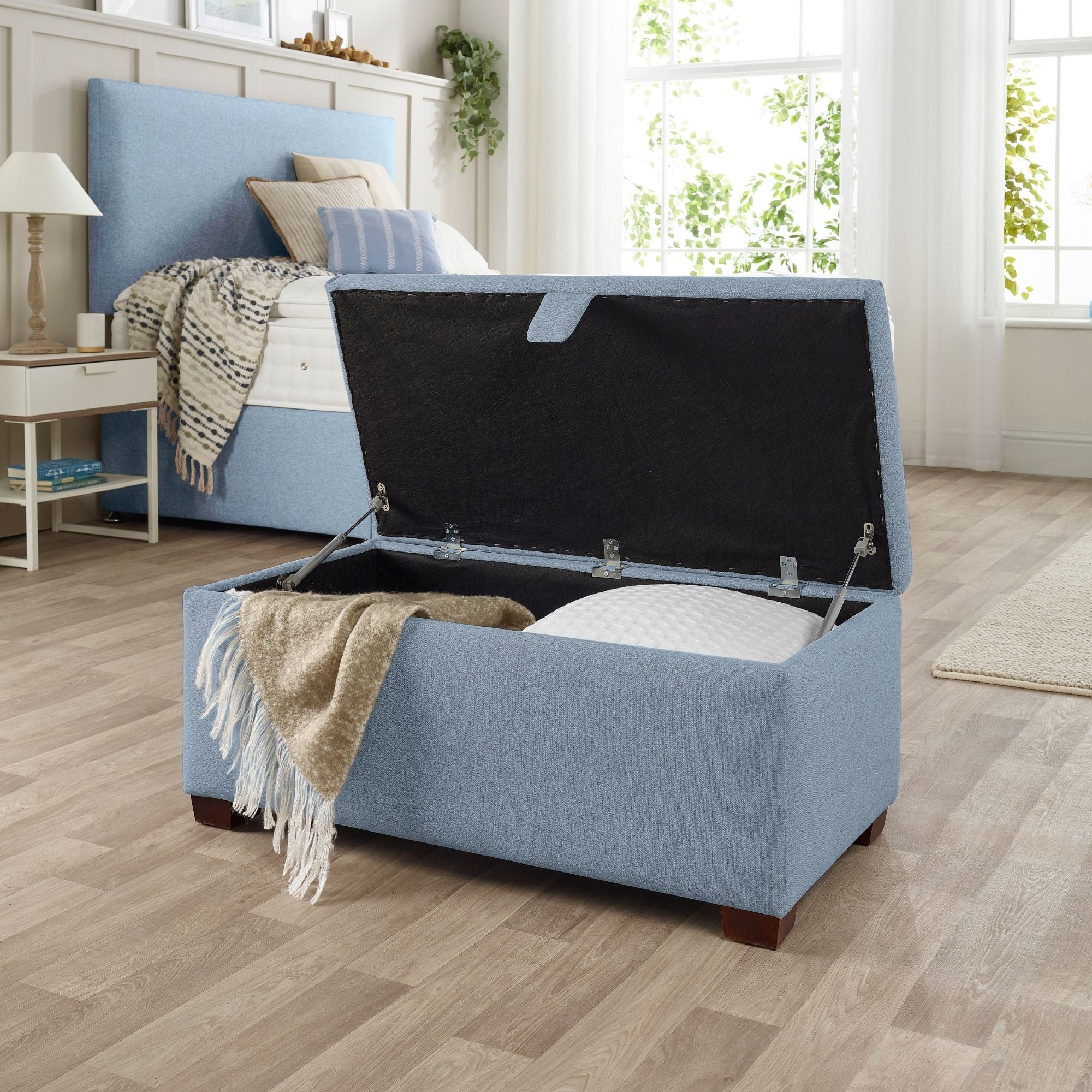 The Woolroom Ottoman Storage Box in Juniper light blue upholstery stores pillows and throws and other items.