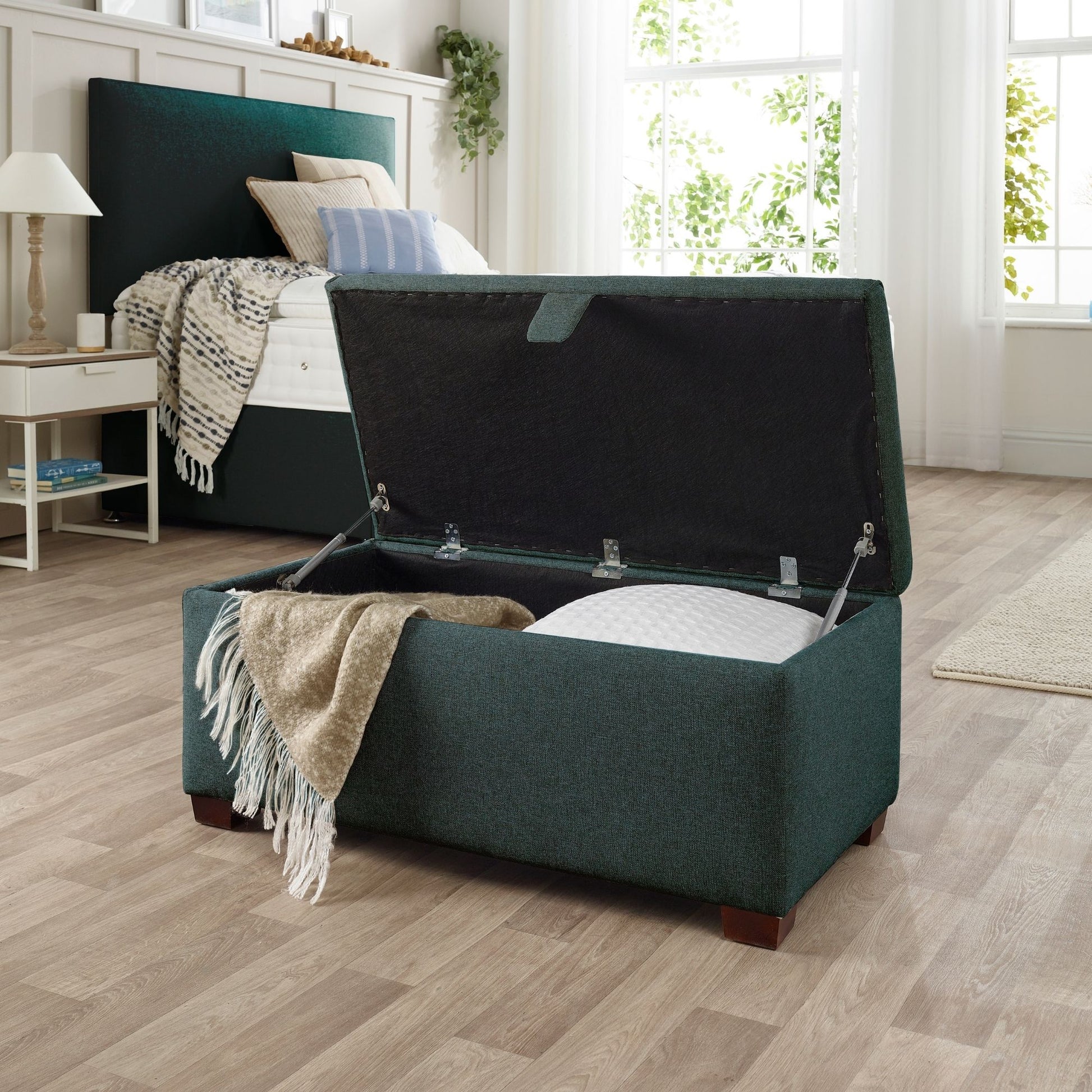 The Woolroom Ottoman Storage Box in Pine dark green upholstery stores pillows and throws and other items.