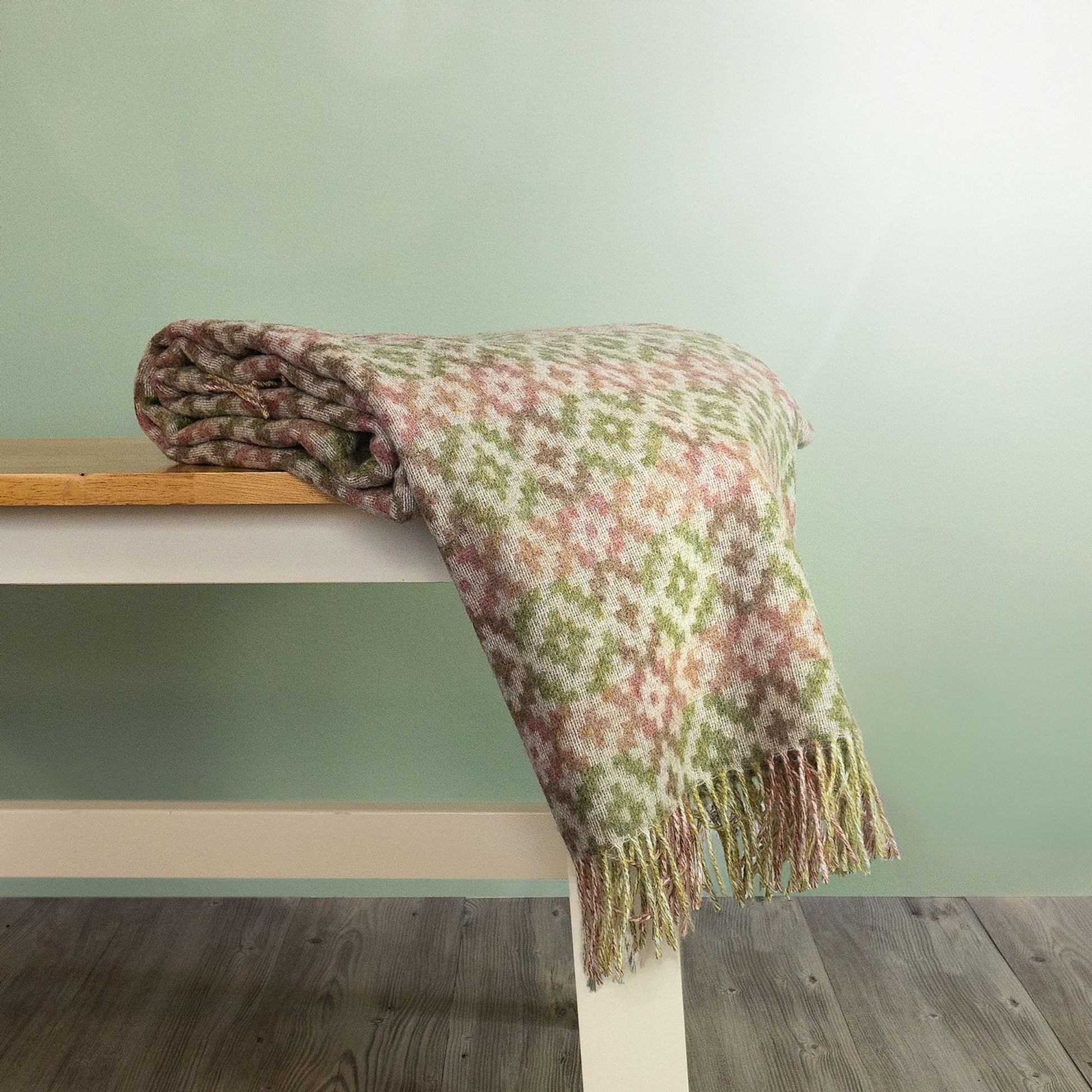The Dartmouth Wool Throw in Green is a unique cross and square pattern and fringe, shown folded on a bench.