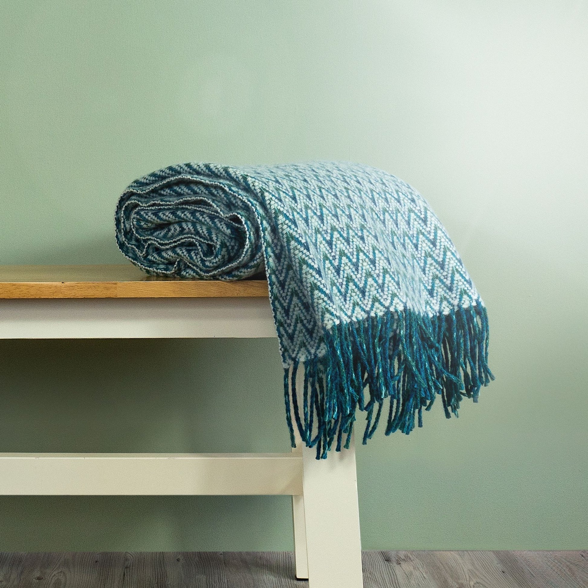The Shady Wave Wool Throw in colour Aqua is a chevron mix of white and teals with fringe, rolled up on a bench.