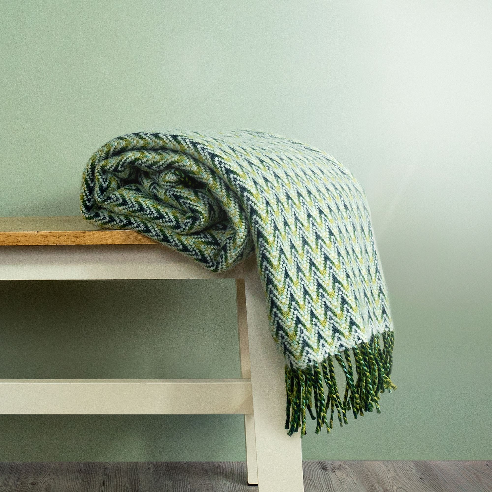 The Shady Wave Wool Throw in colour Green is a chevron mix of white and greens with fringe, rolled up on a bench.