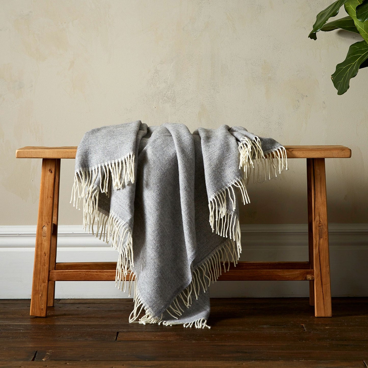 The Woolroom Lilly Herringbone Pure New Wool Throw in Grey has a finer pattern detail and is draped over a bench with fringe.