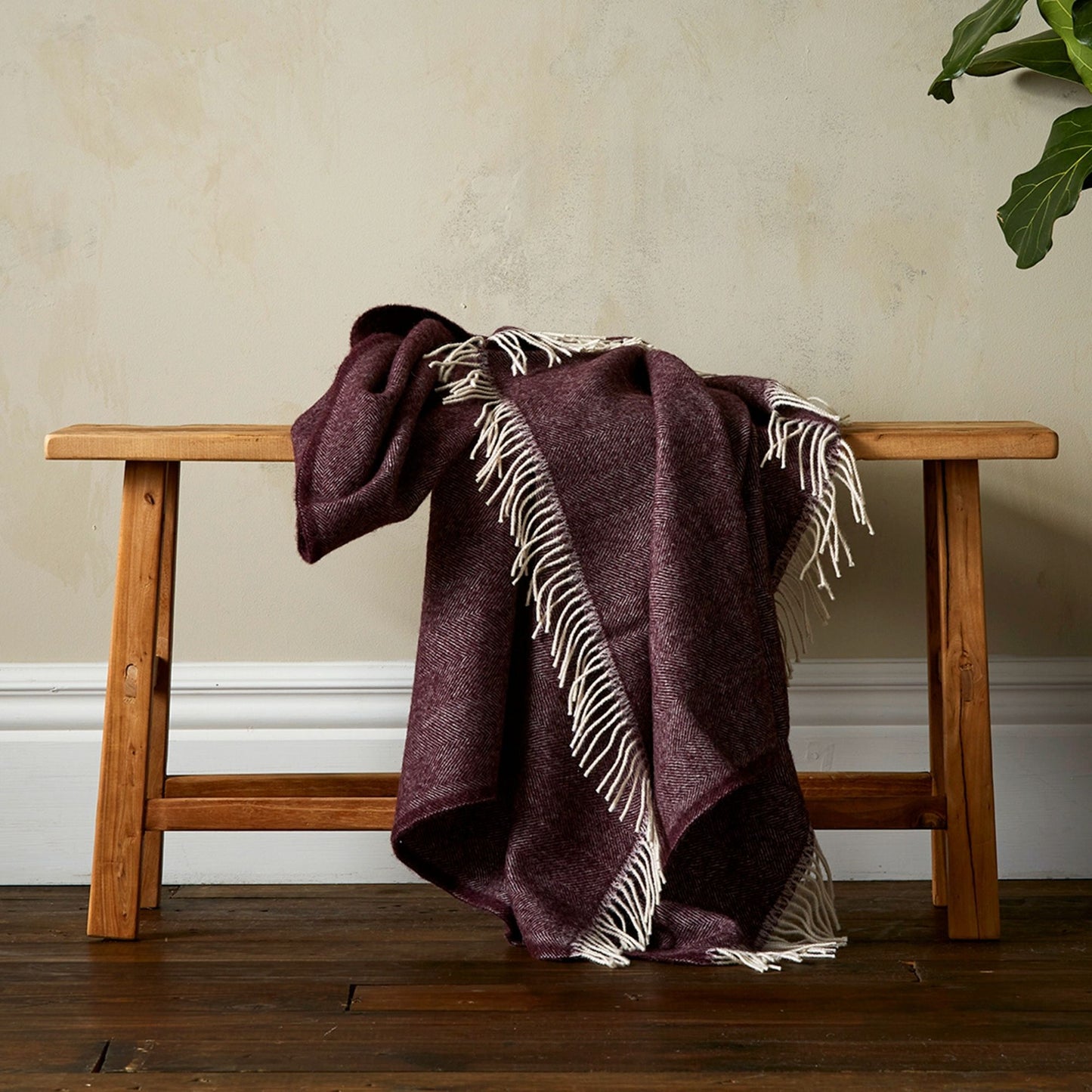 The Woolroom Lilly Herringbone Pure New Wool Throw in Plum has a finer pattern detail and is draped over a bench with fringe.