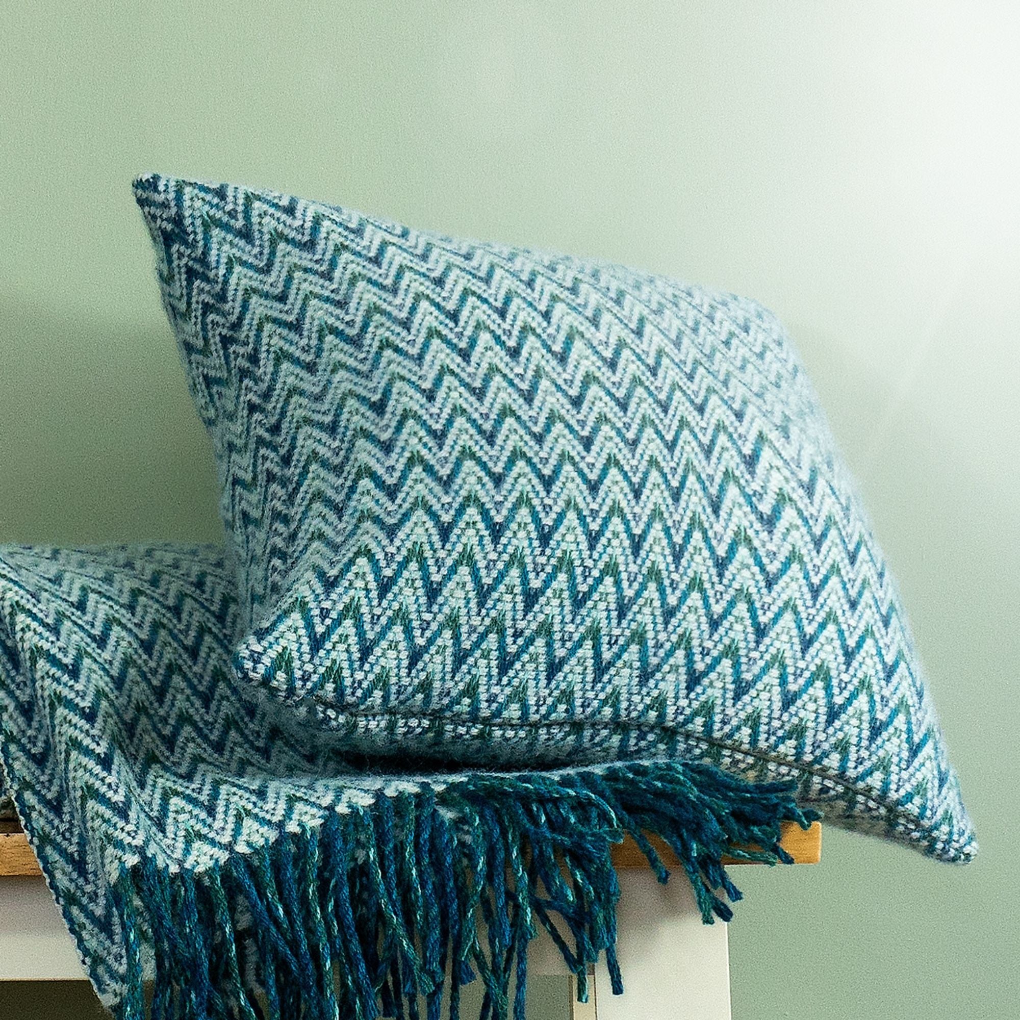 The Shady Wave Wool Cushion Cover in colour Aqua is a chevron mix of white and teals, shown with the matching throw blanket.