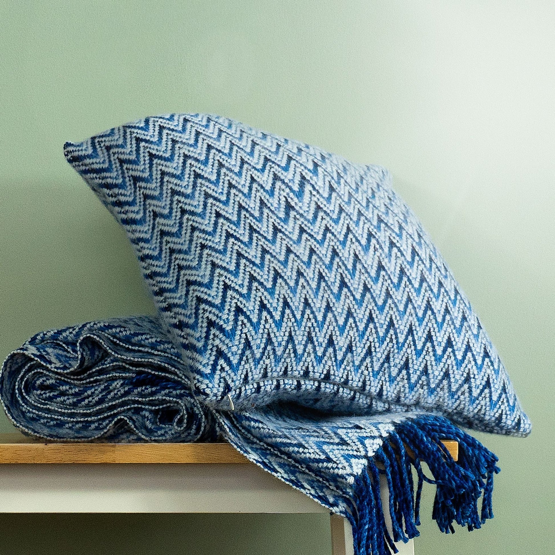 The Shady Wave Wool Cushion Cover in colour Blue is a chevron mix of white and teals, shown with the matching throw blanket.