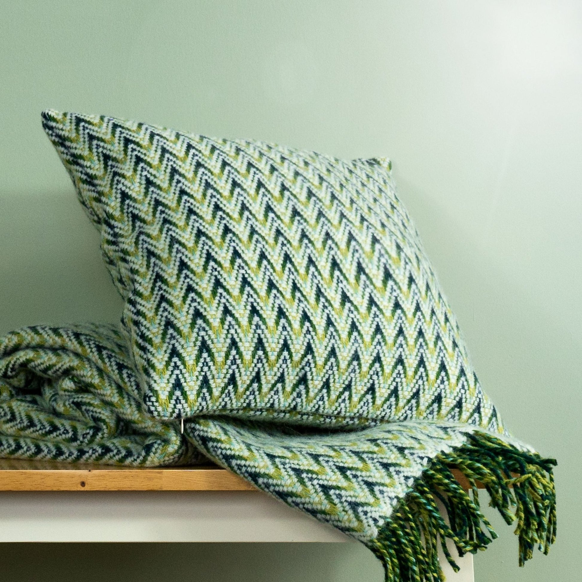 The Shady Wave Wool Cushion Cover in colour Green is a chevron mix of white and teals, shown with the matching throw blanket.