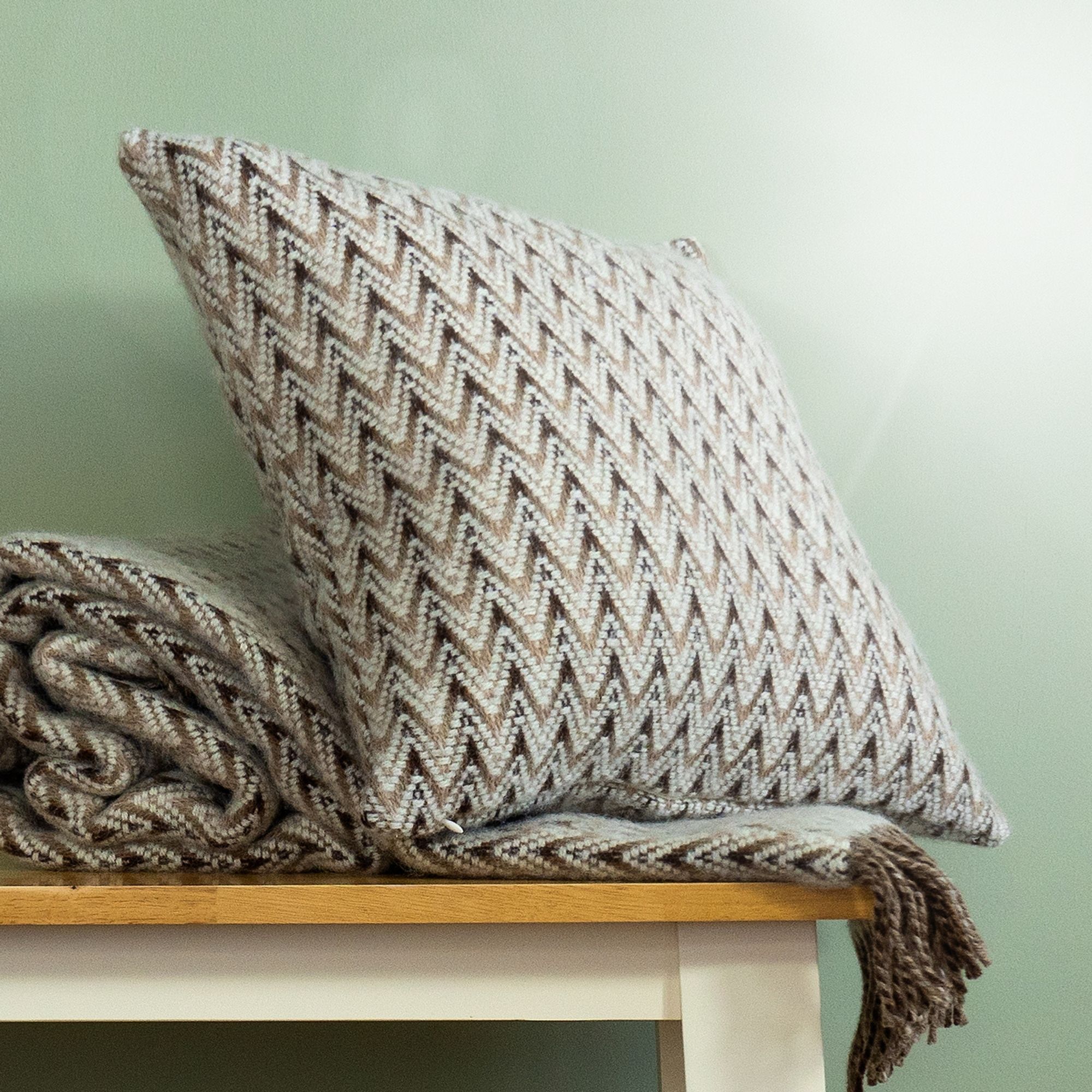 The Shady Wave Wool Cushion Cover in colour Natural is a chevron mix of white and teals, shown with the matching throw blanket.