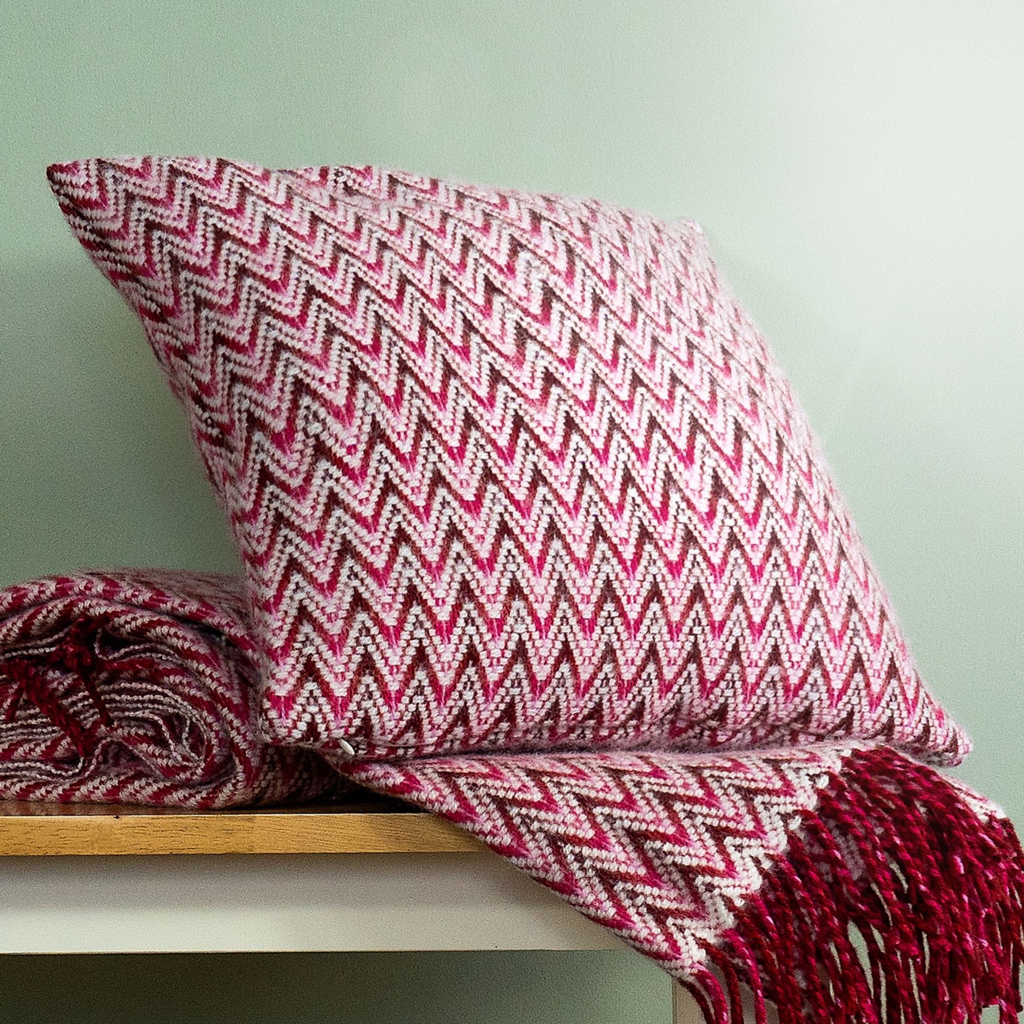 The Shady Wave Wool Cushion Cover in colour Pink is a chevron mix of white and teals, shown with the matching throw blanket.