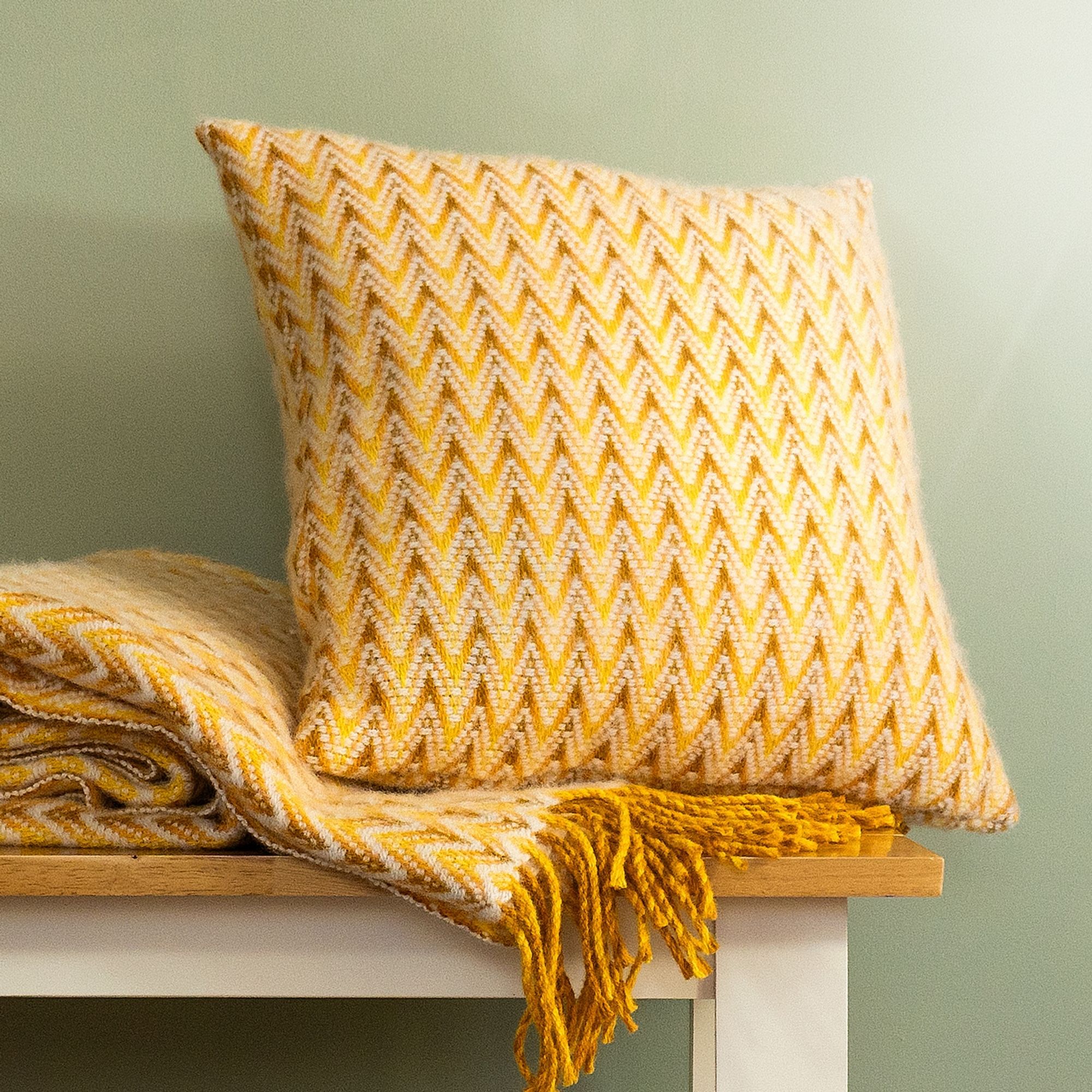 The Shady Wave Wool Cushion Cover in colour Yellow is a chevron mix of white and teals, shown with the matching throw blanket.