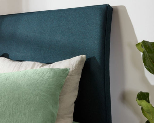 Teal upholstered headboard with pillows and a plant on a light background