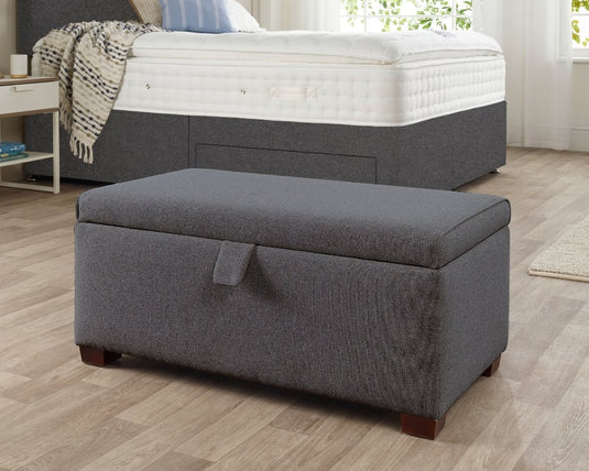 Gray upholstered storage ottoman in a bedroom setting with a bed in the background.