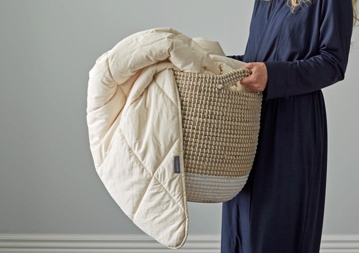 Person holding a woven basket with a beige quilt draped over it against a plain background