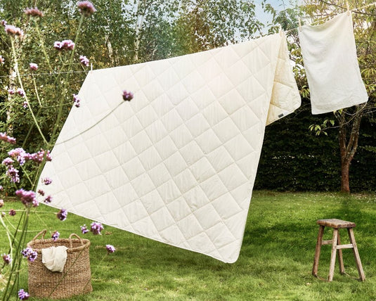 Quilted coverlet hanging outdoors on a line with flowers and a stool in the background