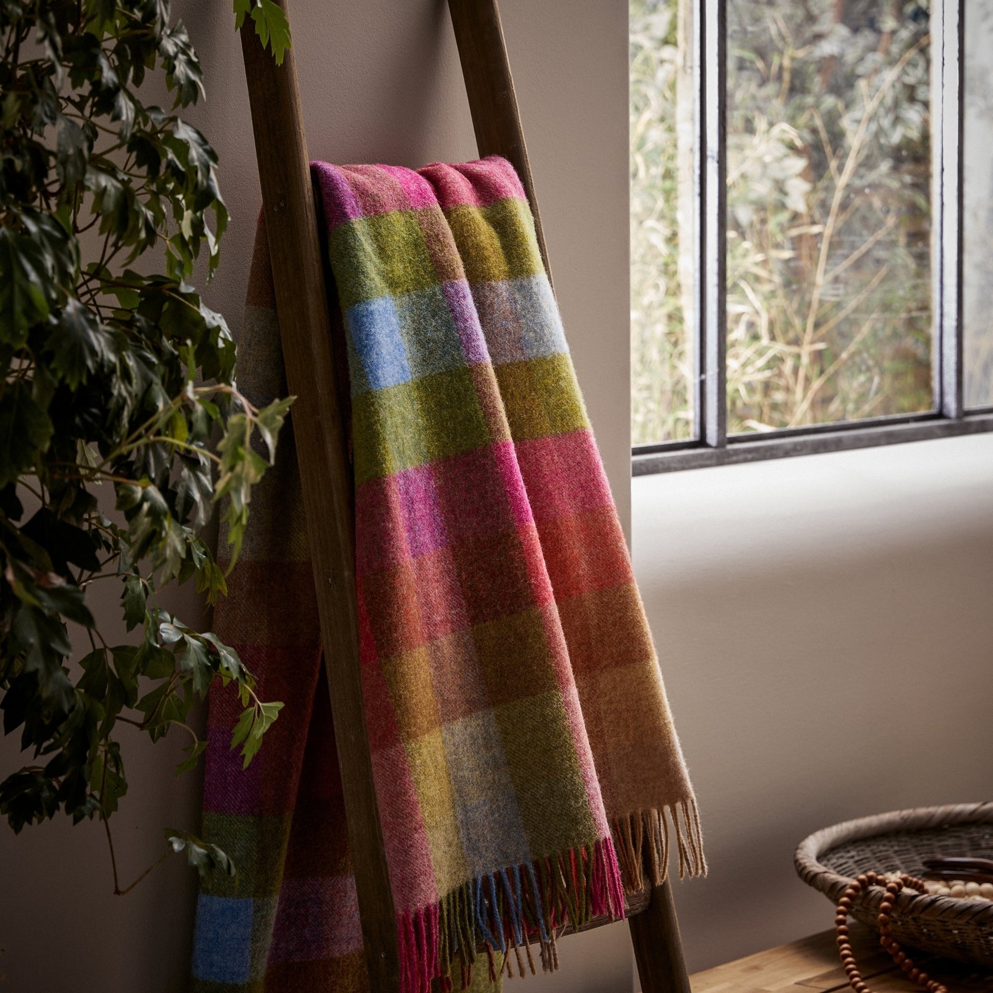 The Harlequin Shetland Wool Throw in color Sunshine is a bright mix of pinks, yellows, greens, and blues, draped over a ladder.