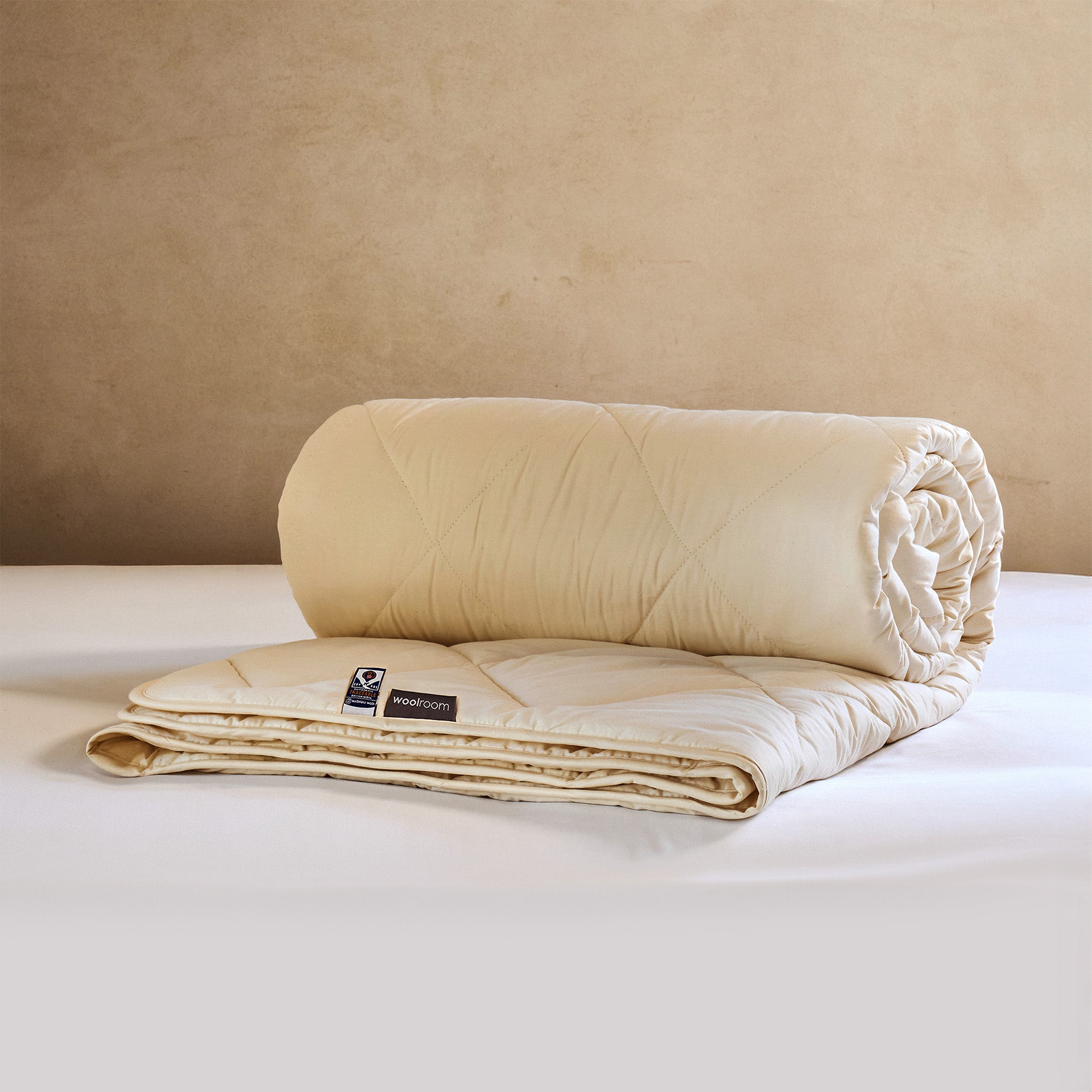 A cream quilted wool duvet, part rolled and part folded on a bed, with a Woolroom label at the hem.
