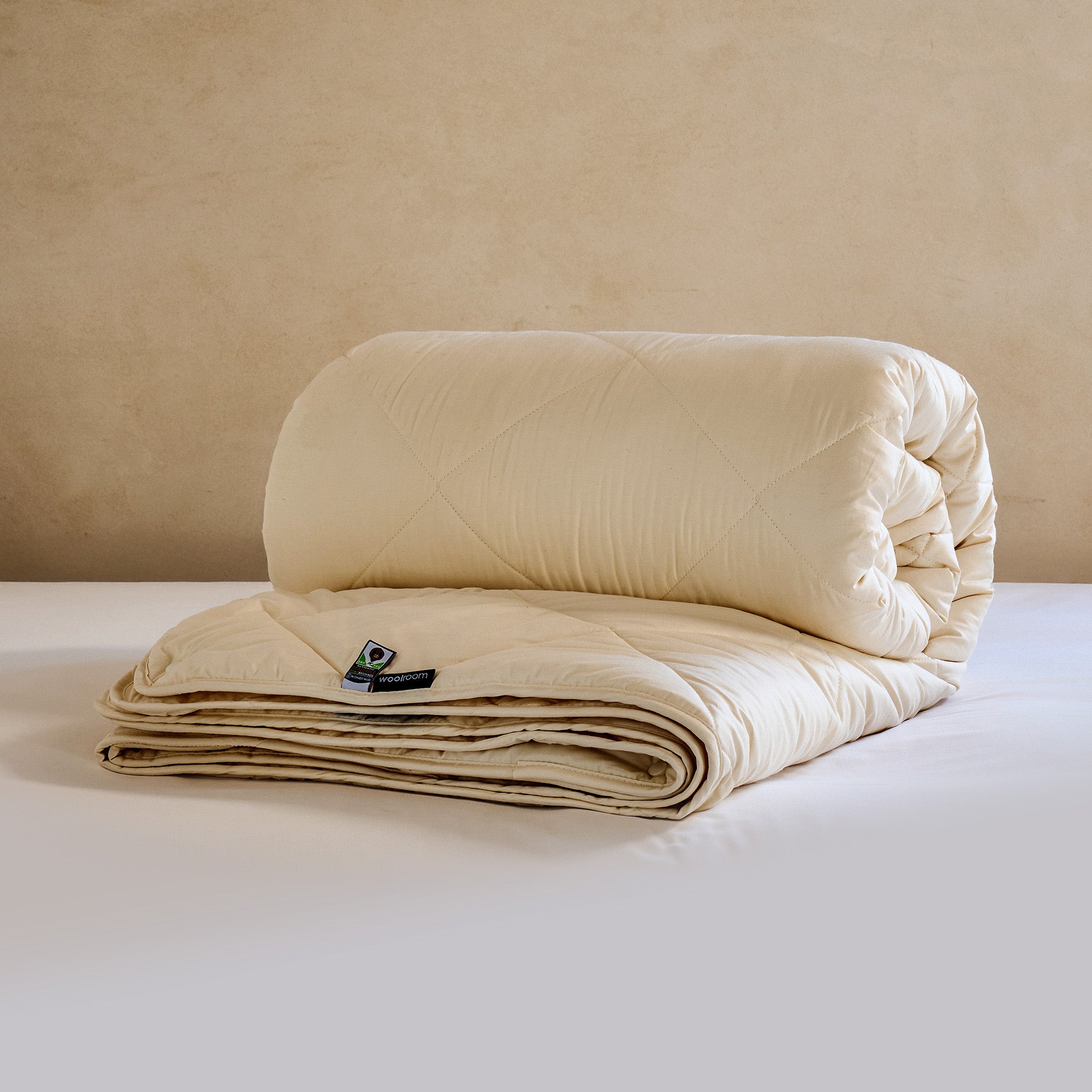 A cream quilted organic wool duvet shown rolled on a bed, with a Woolroom label at the hem.