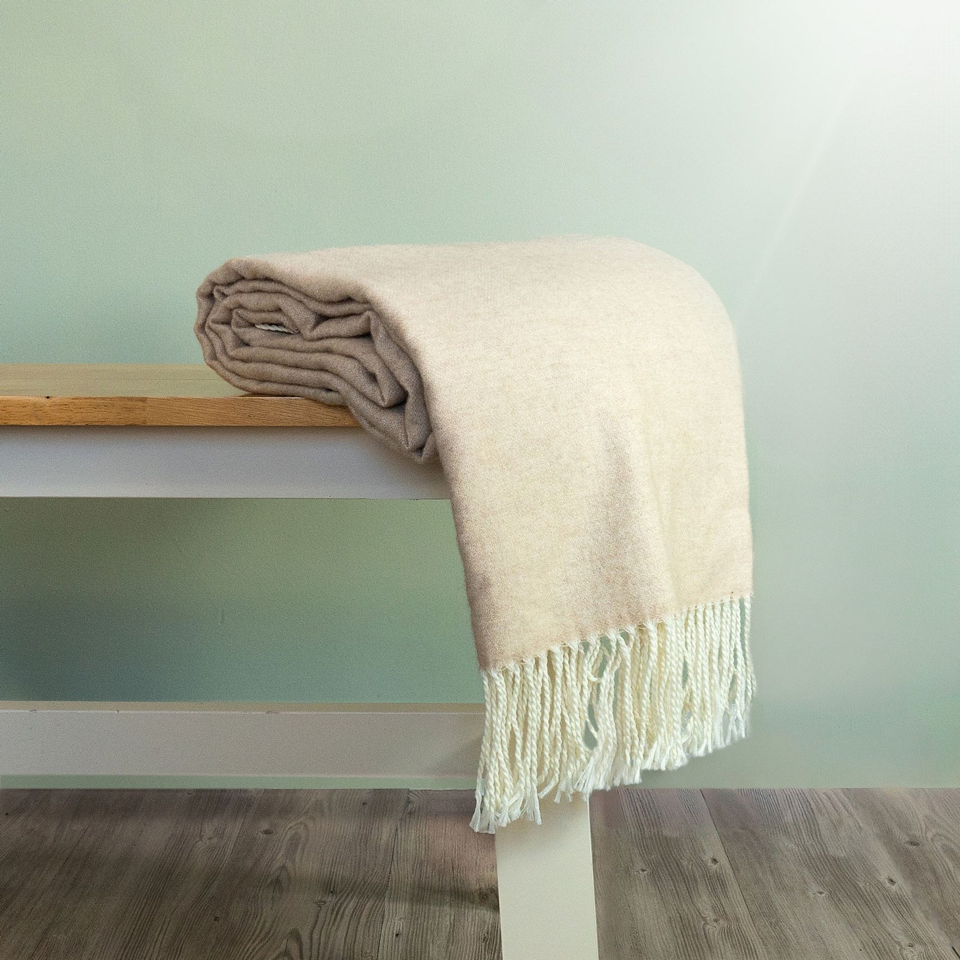The Como Wide Wool Throw in light grey with beige fringe on the ends is rolled up and placed on a bench.