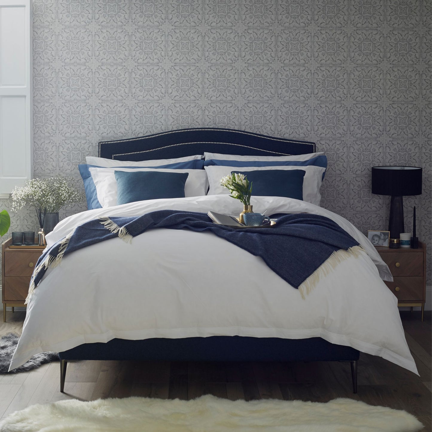 The Olivia Upholstered Bedstead shown in a navy blue with nailhead trim around the curved headboard with a fully dressed bed.