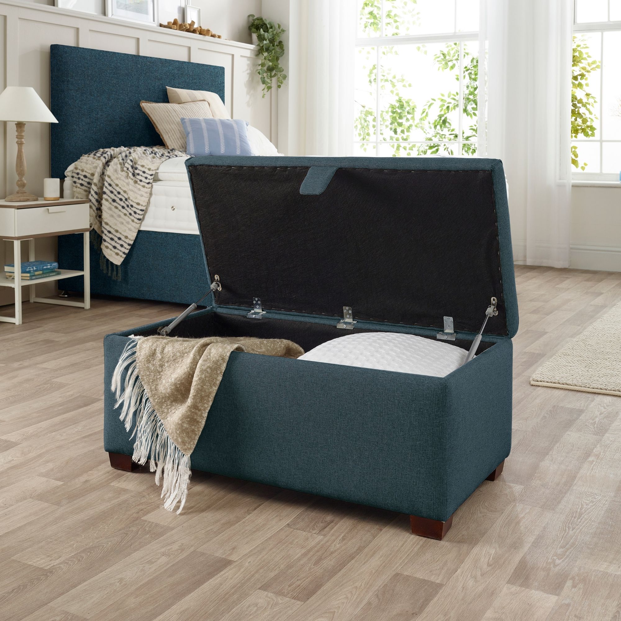 The Woolroom Ottoman Storage Box in Hemlock blue upholstery stores pillows and throws and other items. 