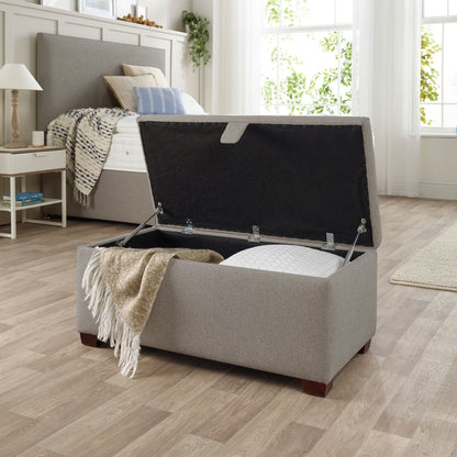 The Woolroom Ottoman Storage Box in light grey upholstery stores pillows and throws and other items. 