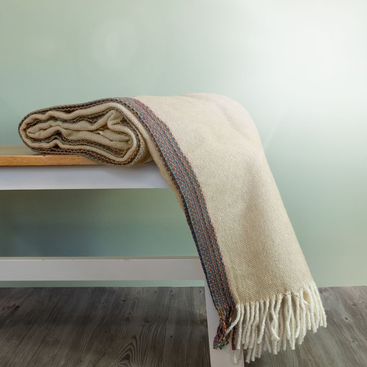 The Aurora Wool Throw in colour Beige has a multi-coloured woven edging, shown draped on a bench with cream fringe. 