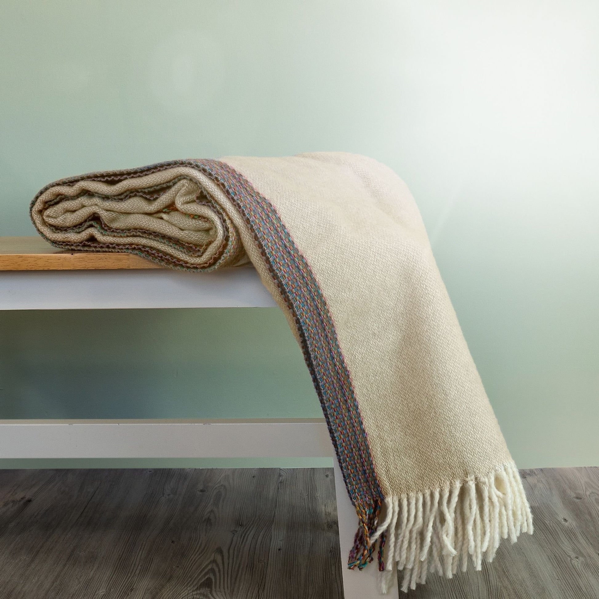 The Aurora Wool Throw in colour Beige has a multi-coloured woven edging, shown draped on a bench with cream fringe. 