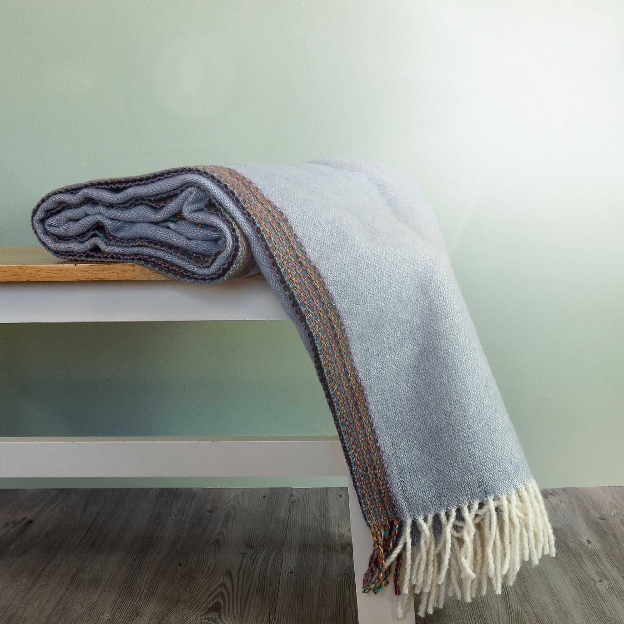 The Aurora Wool Throw in colour Grey has a multi-coloured woven edging, shown draped on a bench with cream fringe. 