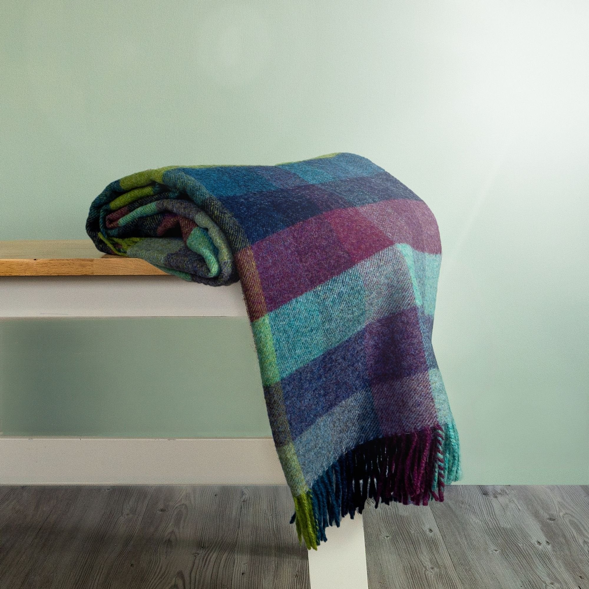The Moorland Wool Throw in colour Harlequin is a cool-toned mix of dark purple, blue, green, rolled on a bench showing the fringe. 