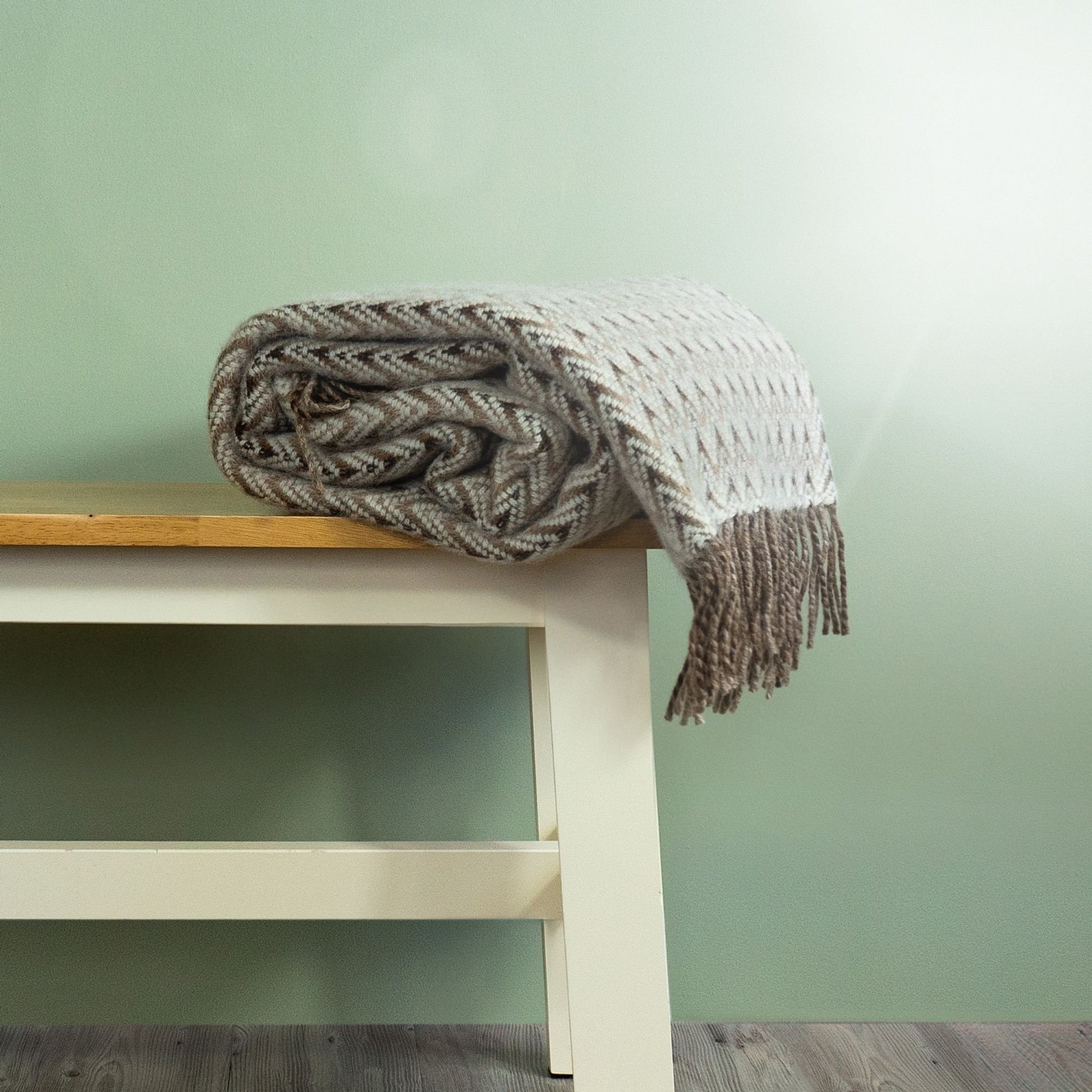 The Shady Wave Wool Throw in colour Natural is a chevron mix of white and greens with fringe, rolled up on a bench. 