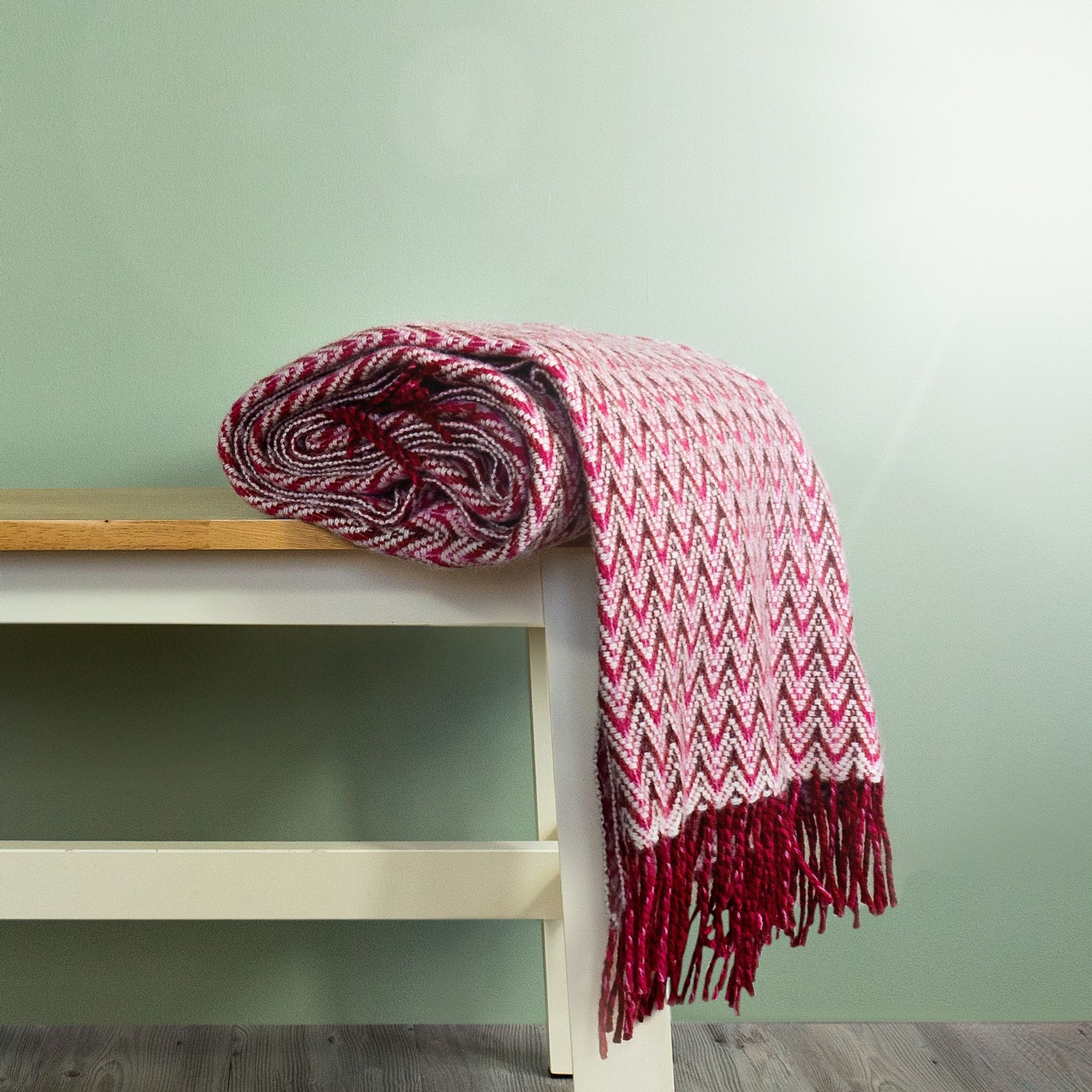 The Shady Wave Wool Throw in colour Pink is a chevron mix of white and pinks with fringe, rolled up on a bench. 