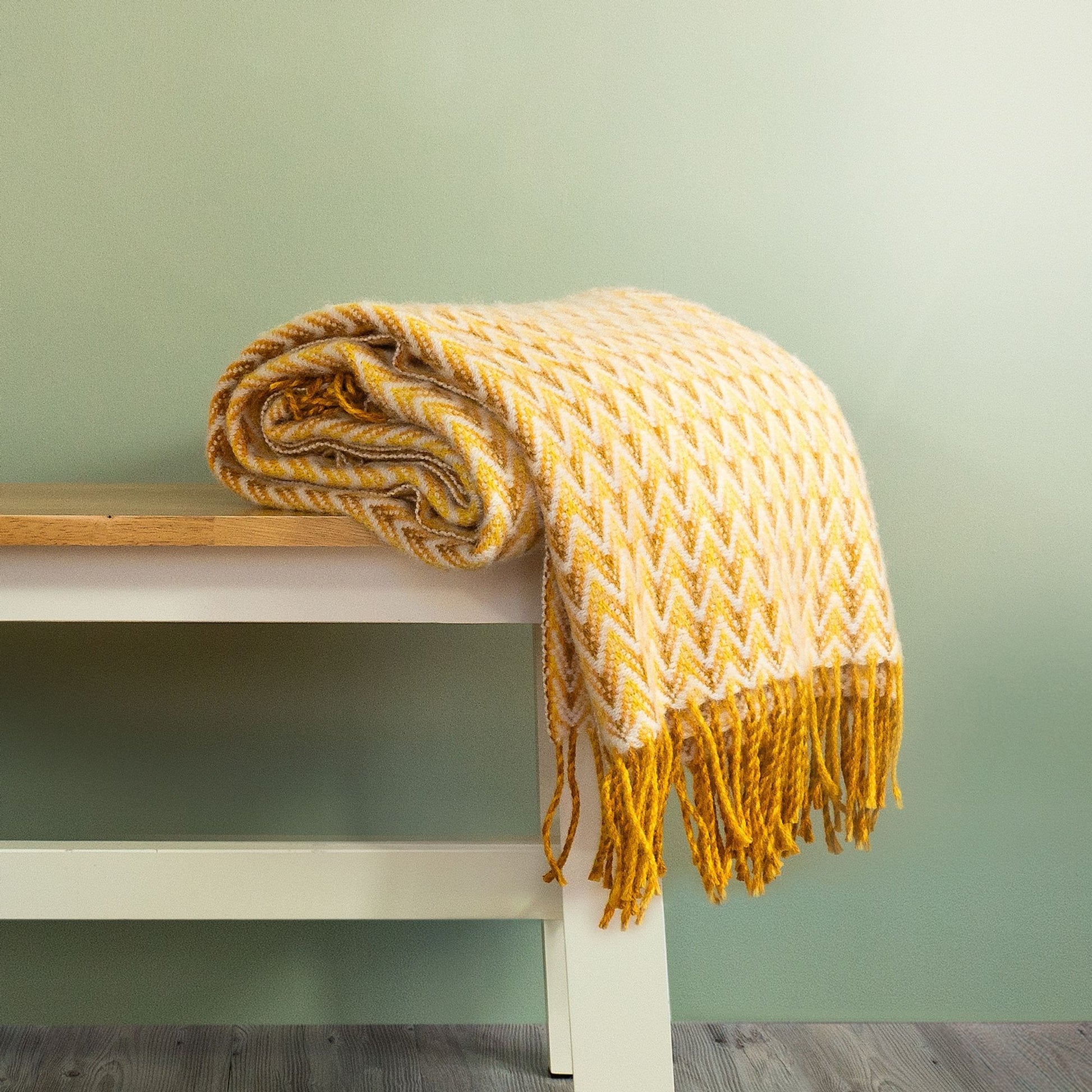 The Shady Wave Wool Throw in colour Yellow is a chevron mix of white and yellows with fringe, rolled up on a bench. 