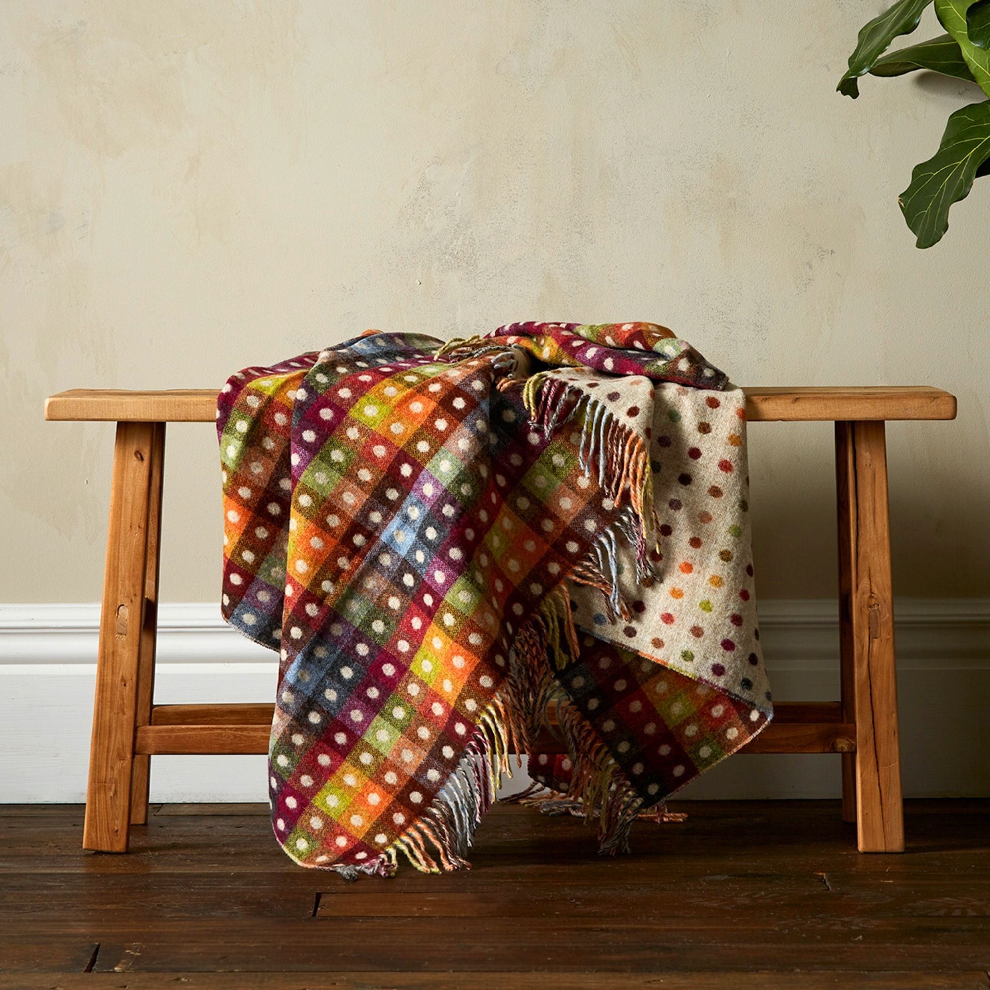 The Beige Rainbow Spot Throw is a bright mix of colours in a block and dot pattern, draped over a bench showing the fringe. 