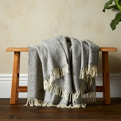 The super soft Greta Herringbone Wool Blanket in Grey draped over a bench with white fringe. 