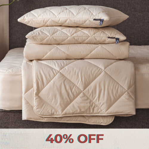 A blonde lady sitting on a mattress, with a pile of Woolroom bedding, including a duvet, pillows and mattress protector