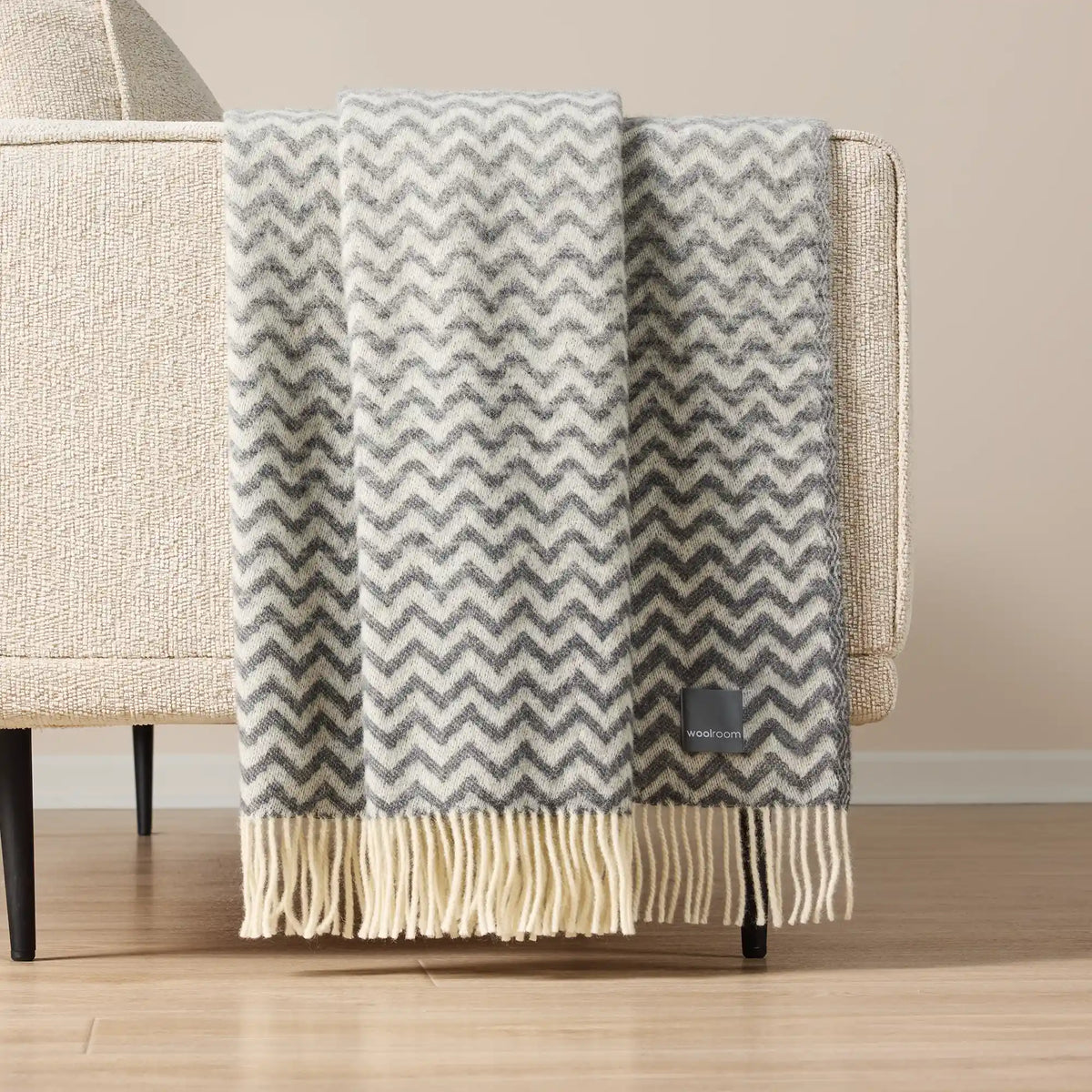 A gray and cream chevron pattern throw blanket with fringe, draped over the arm of a beige armchair in a minimalist room setting.