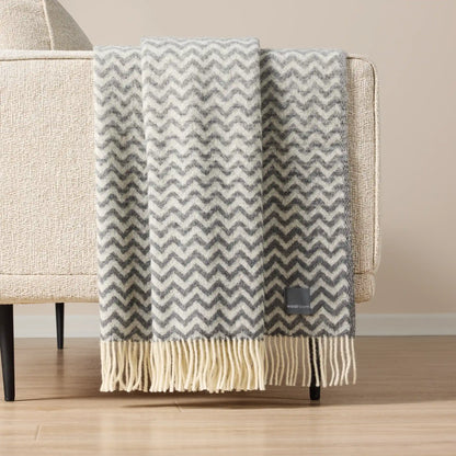 A gray and cream chevron pattern throw blanket with fringe, draped over the arm of a beige armchair in a minimalist room setting.