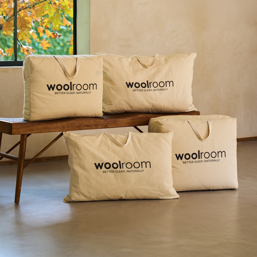 Une dame blonde assise sur un matelas, avec une pile de literie Woolroom, comprenant une couette, des oreillers et un protège-matelas