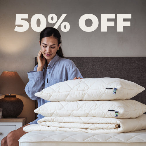 A blonde lady sitting on a mattress, with a pile of Woolroom bedding, including a duvet, pillows and mattress protector