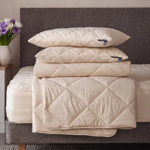 A blonde lady sitting on a mattress, with a pile of Woolroom bedding, including a duvet, pillows and mattress protector