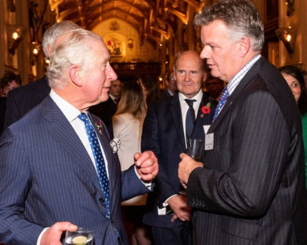 Chris Tattersall and King Charles in dark pinstripe suits shaking hands in a formal indoor event setting with a warm, ornate background.