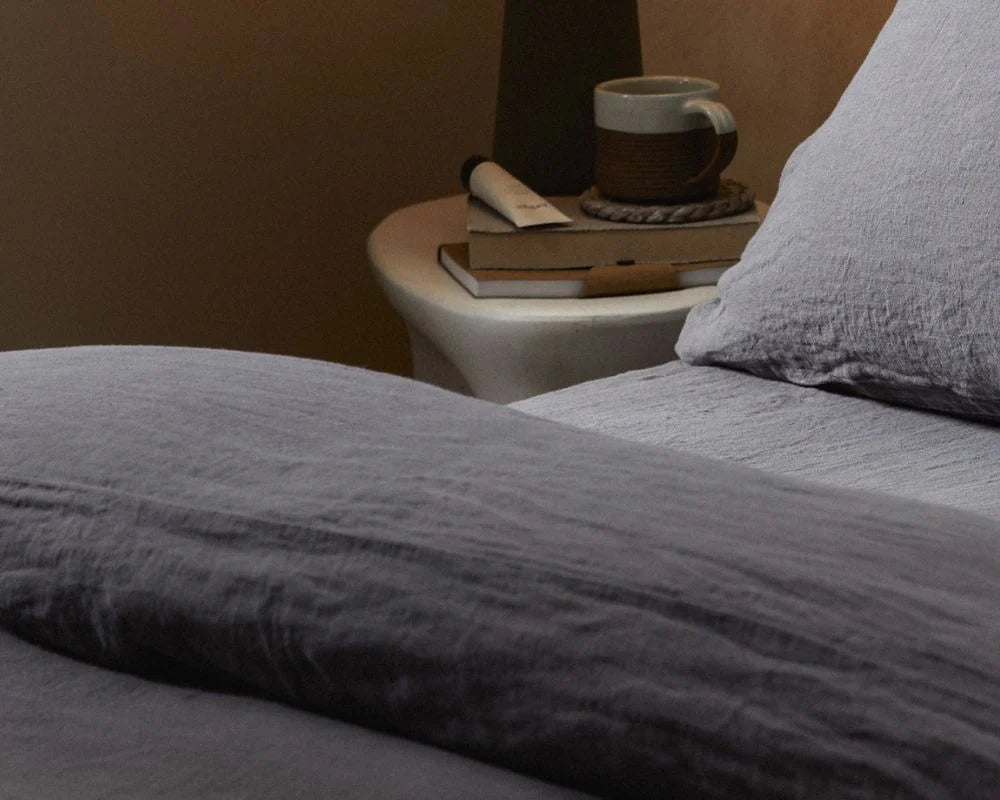 Gray bedding on a bed with a side table featuring a mug and books in the background.