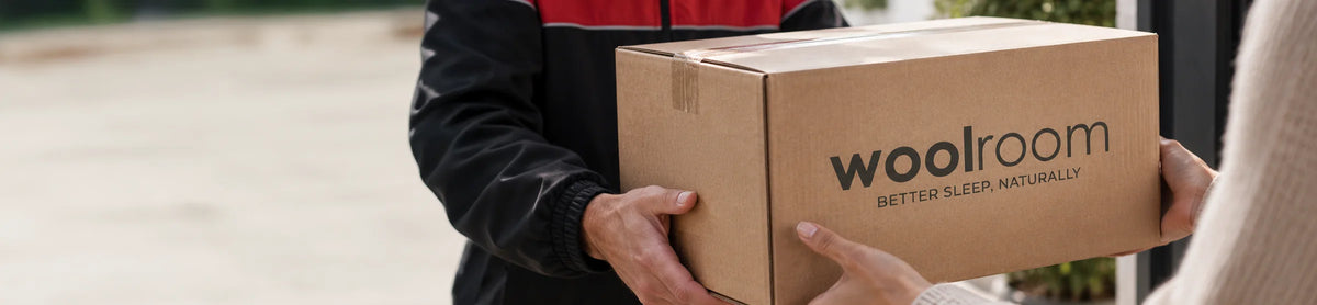 A person in a black and red jacket holds a brown cardboard box printed with the brand name 'woolroom' and the tagline 'BETTER SLEEP NATURALLY' while being handed to another person.
