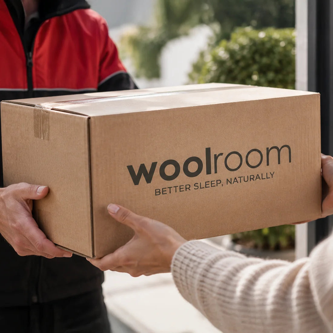 A person in a red and black uniform holds a large brown cardboard box printed with the brand name 'woolroom' and the tagline 'BETTER SLEEP, NATURALLY' while being handed by another person in a beige sweater.