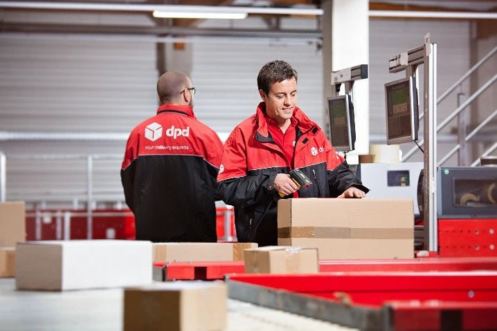 Two DPD delivery workers in red and black uniforms handling cardboard boxes on a conveyor belt inside a warehouse.