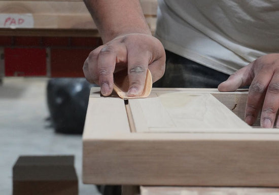 A close-up of a person's hands using a sanding block to smooth the edge of a light-colored wooden cabinet or frame in a workshop setting.