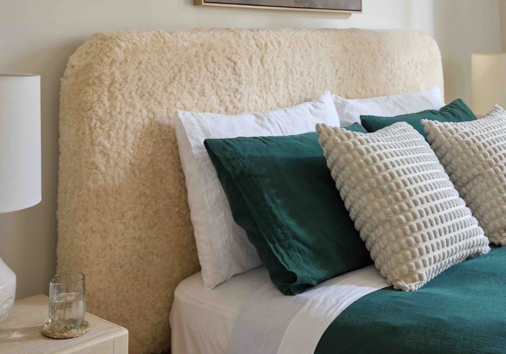 A close-up of a bed with a large, shaggy beige headboard, styled with white and dark green pillows and a green throw blanket. A white bedside lamp and a small glass on a coaster are visible on the left side.