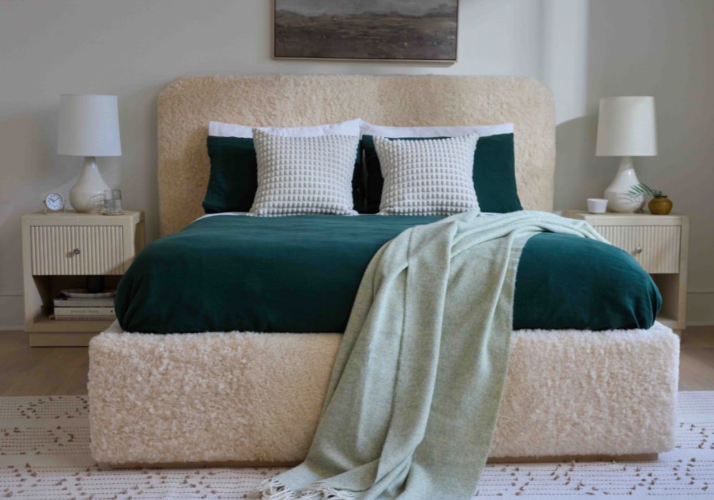 A modern platform bed with a textured cream boucle headboard and base, dressed in dark green velvet bedding and a light sage green throw blanket, flanked by matching nightstands with white lamps.