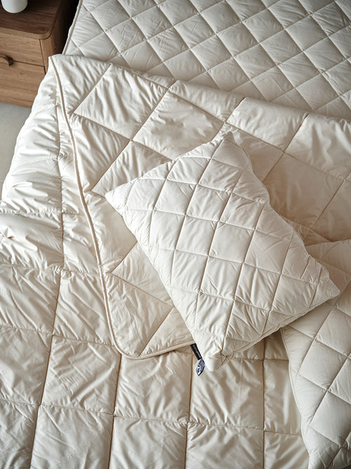 A blonde lady sitting on a mattress, with a pile of Woolroom bedding, including a duvet, pillows and mattress protector