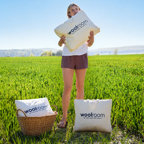 A blonde lady sitting on a mattress, with a pile of Woolroom bedding, including a duvet, pillows and mattress protector